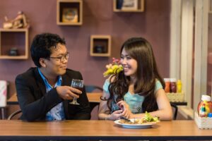 Eating on a date with ARFID [Image description: a man and a woman smiling at each other seated at a table. The man holds a glass and the woman has food in front of her] Depicts a potential couple on a date where one of the members has ARFID